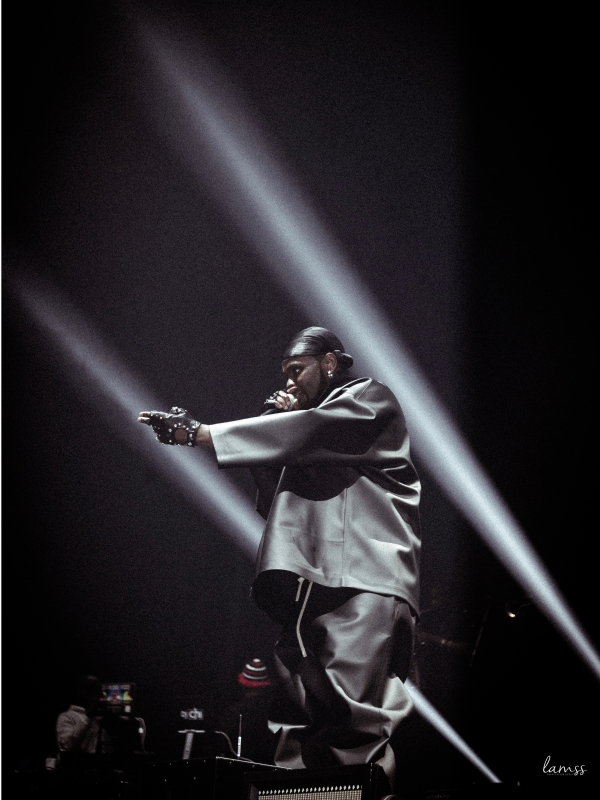 Kizz Daniel in an oversized jacket gesturing toward the crowd under sharp diagonal stage lights. — concert photography London by Lamss Media